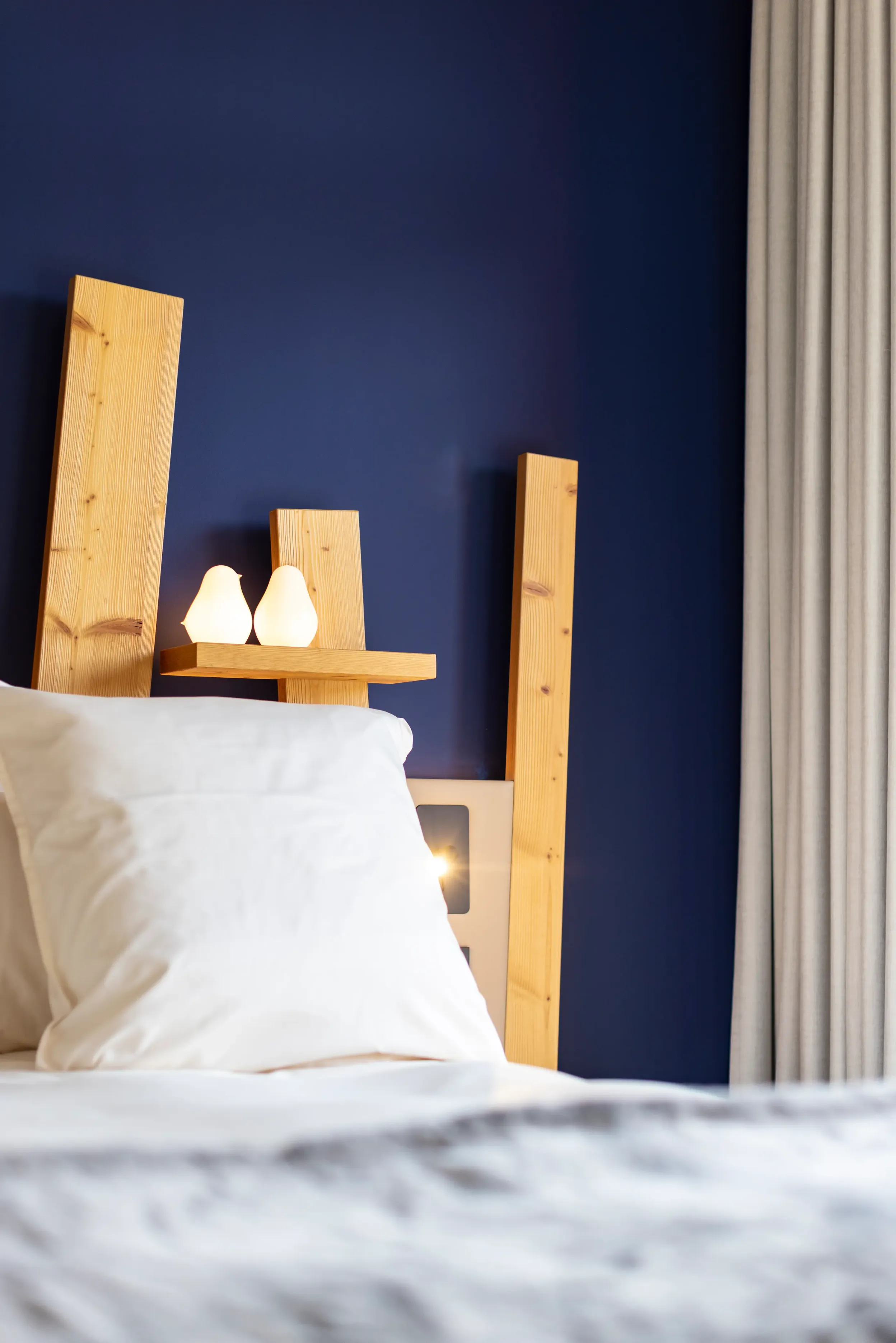 Ein Bett mit einem hellen Holzkopfteil und zwei brennenden Kerzen auf einem Holzregal an einer dunkelblauen Wand