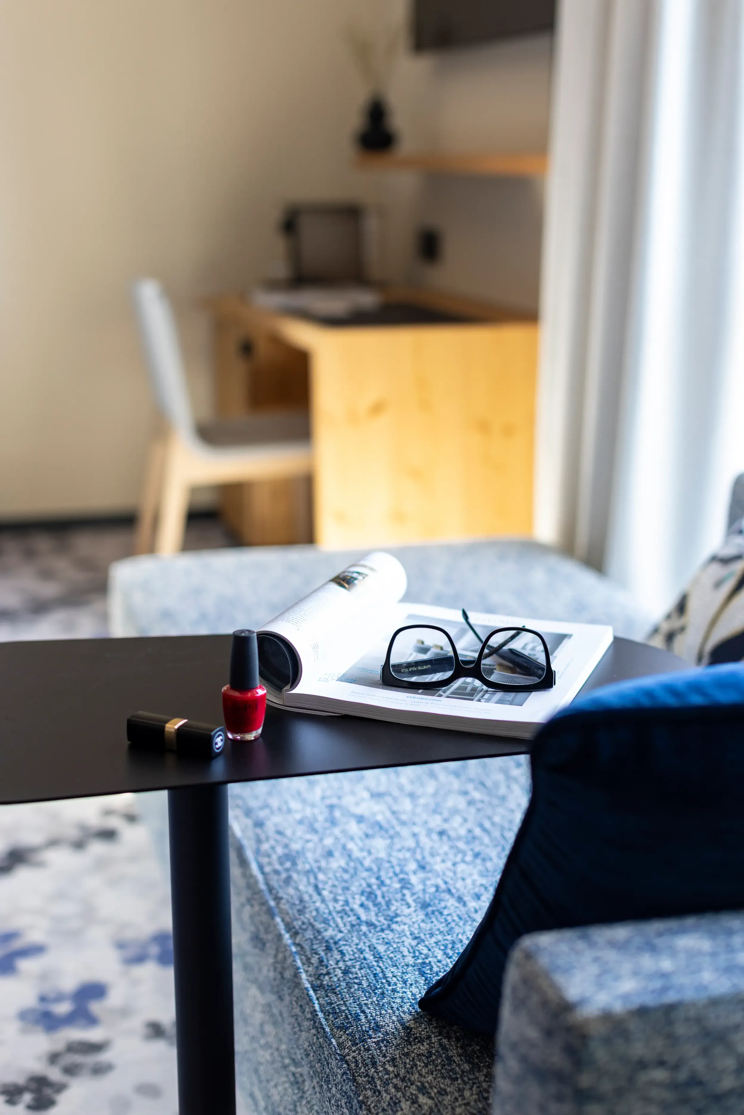 Ein Couchtisch mit einer Flasche roten Nagellacks, einer Brille und einer aufgeschlagenen Zeitschrift, in einem Wohnzimmer mit einem Sofa und einem Holztisch im Hintergrund.