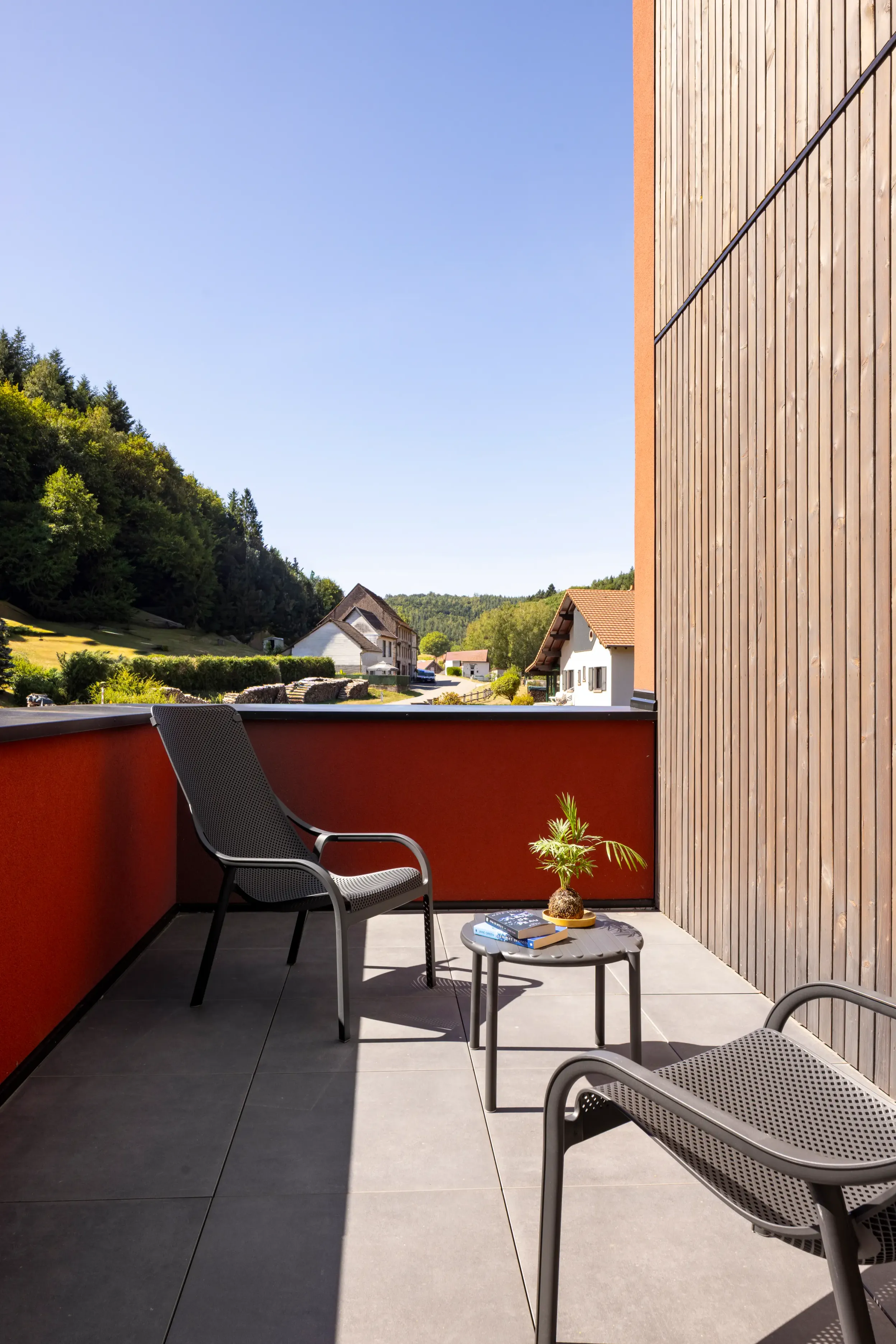 Balcon avec trois chaises et une table basse, vue sur des maisons et des collines boisées