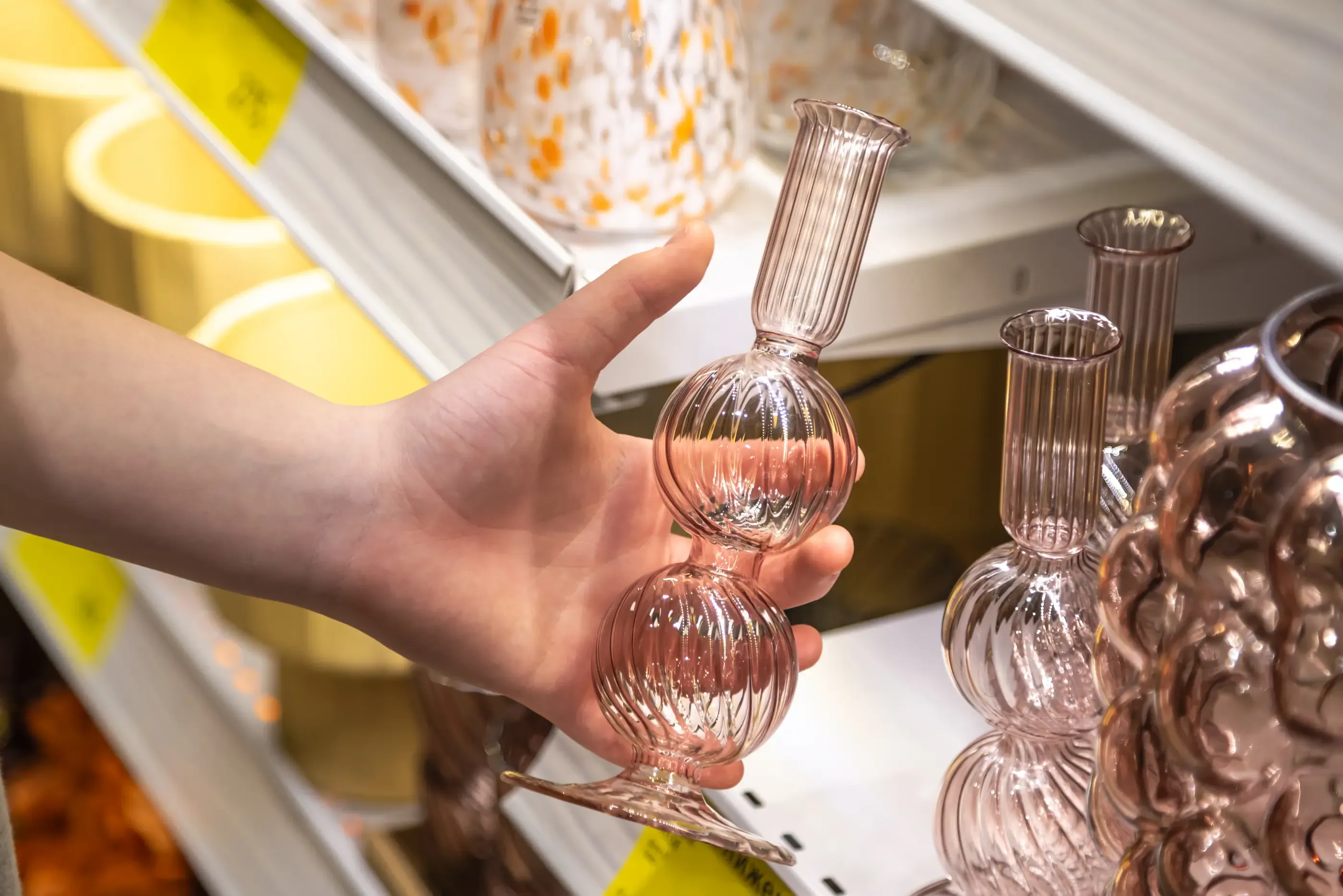 Une main tient un vase en verre soufflé à deux sphères sur une étagère remplie de vases similaires
