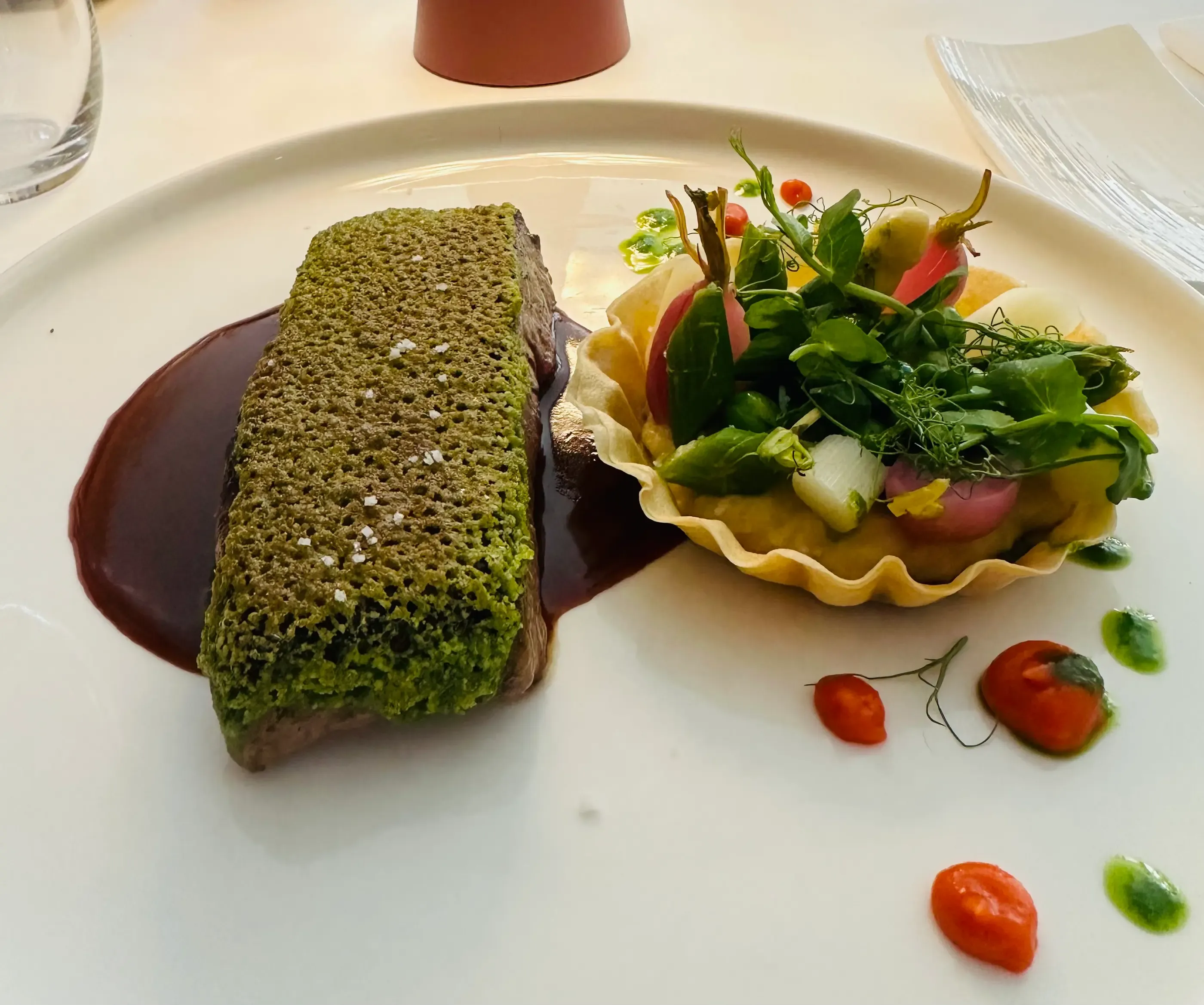 A dish consisting of a crispy green rectangle on a brown sauce, served with a salad of young shoots and cherry tomatoes.