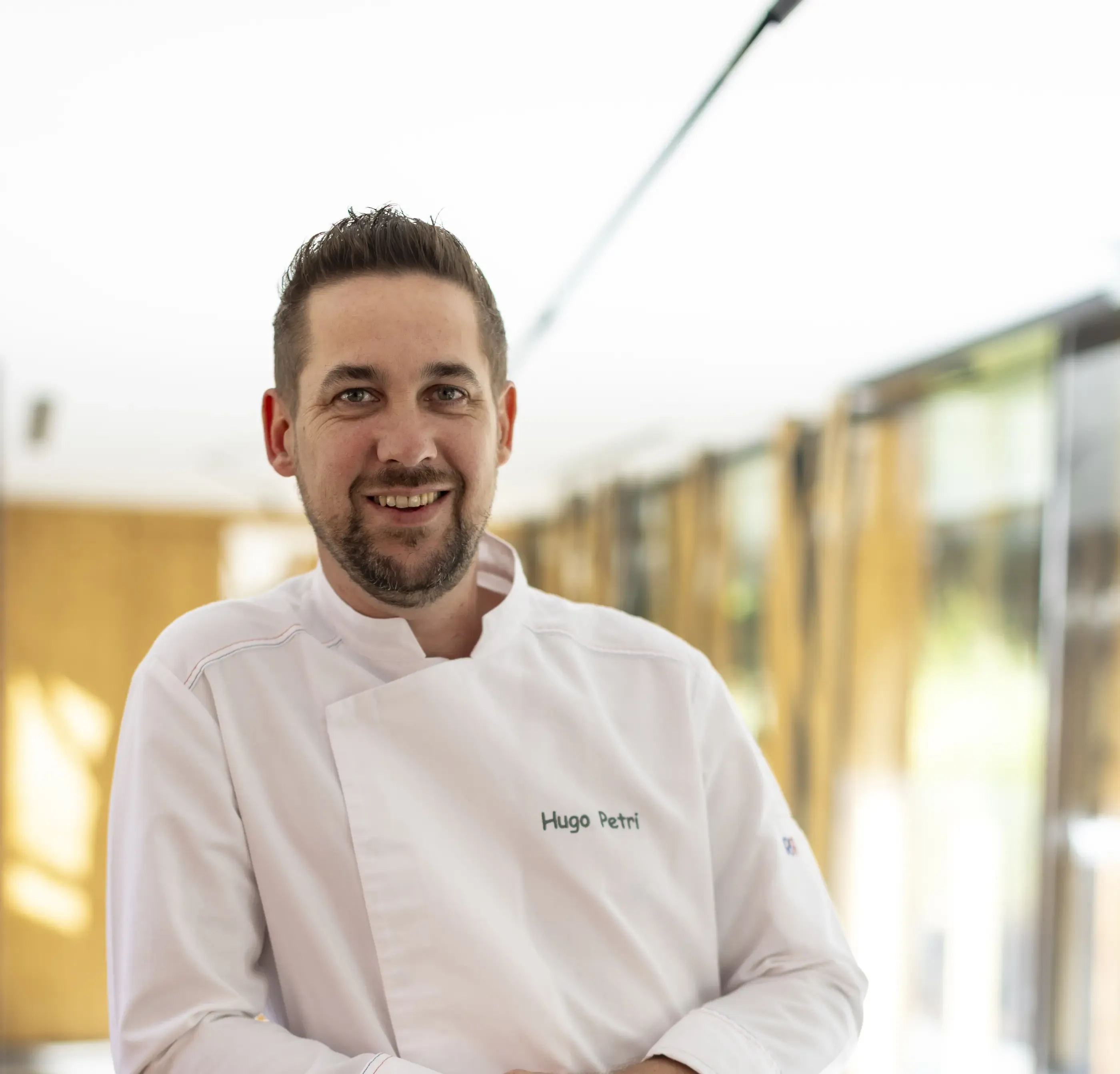 Un homme souriant portant une veste de chef avec le nom "Hugo Petri" brodé dessus, devant un bâtiment vitré