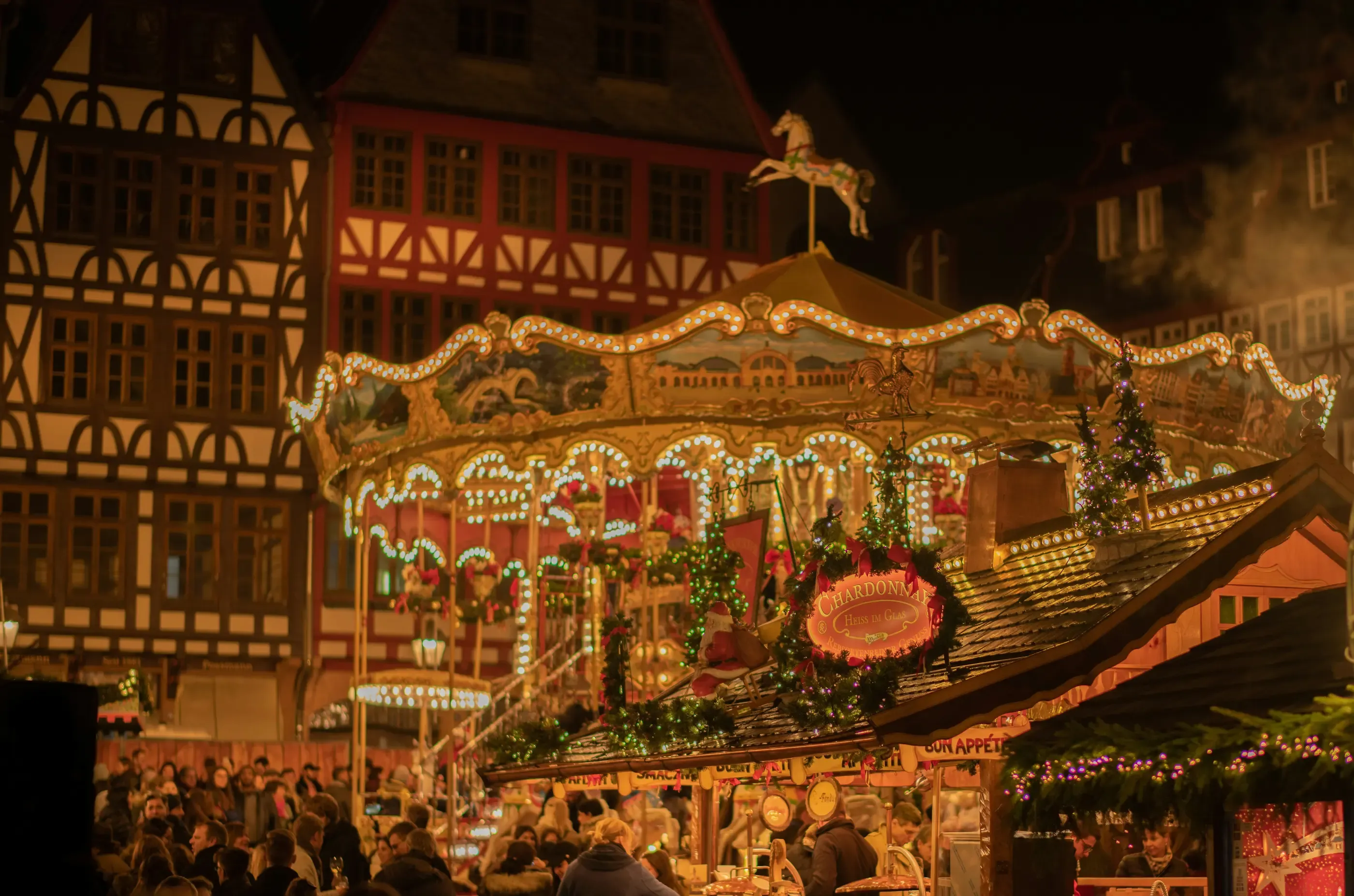 Une grande roue illuminée et décorée de guirlandes est installée sur une place de marché animée, entourée de bâtiments traditionnels à colombages