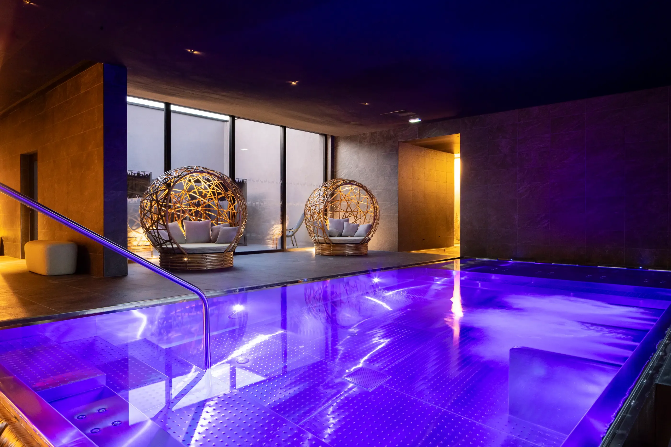 Indoor pool lit in purple with wicker spheres as seats in a modern space with dark walls