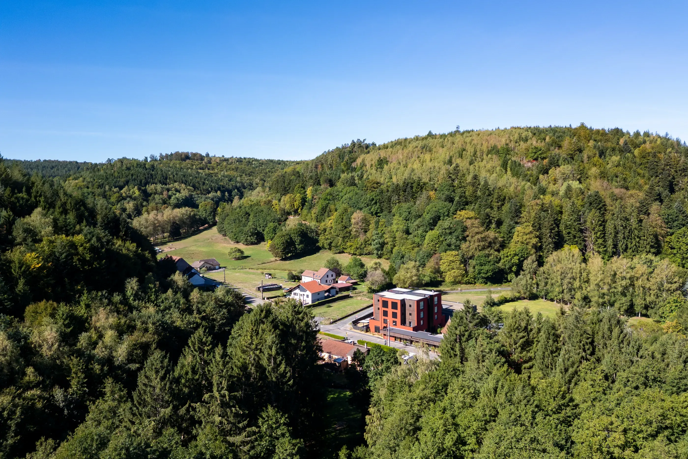 A modern red building surrounded by traditional houses in a wooded valley with a green hill in the background.