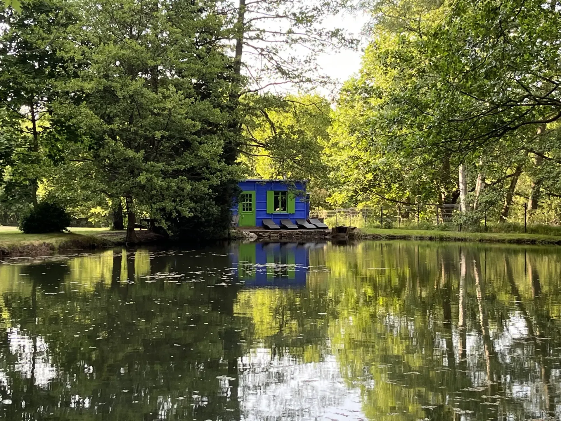 Ein kleines blaues Haus mit grünen Fensterläden steht am Rand eines von Bäumen umgebenen Teichs.