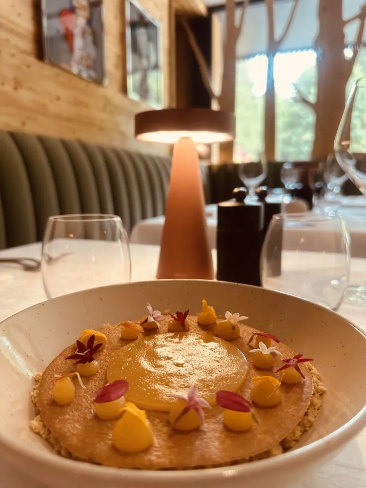 A dish with a circular culinary preparation, garnished with small yellow elements and edible flowers, placed on a table with glasses and a white tablecloth.