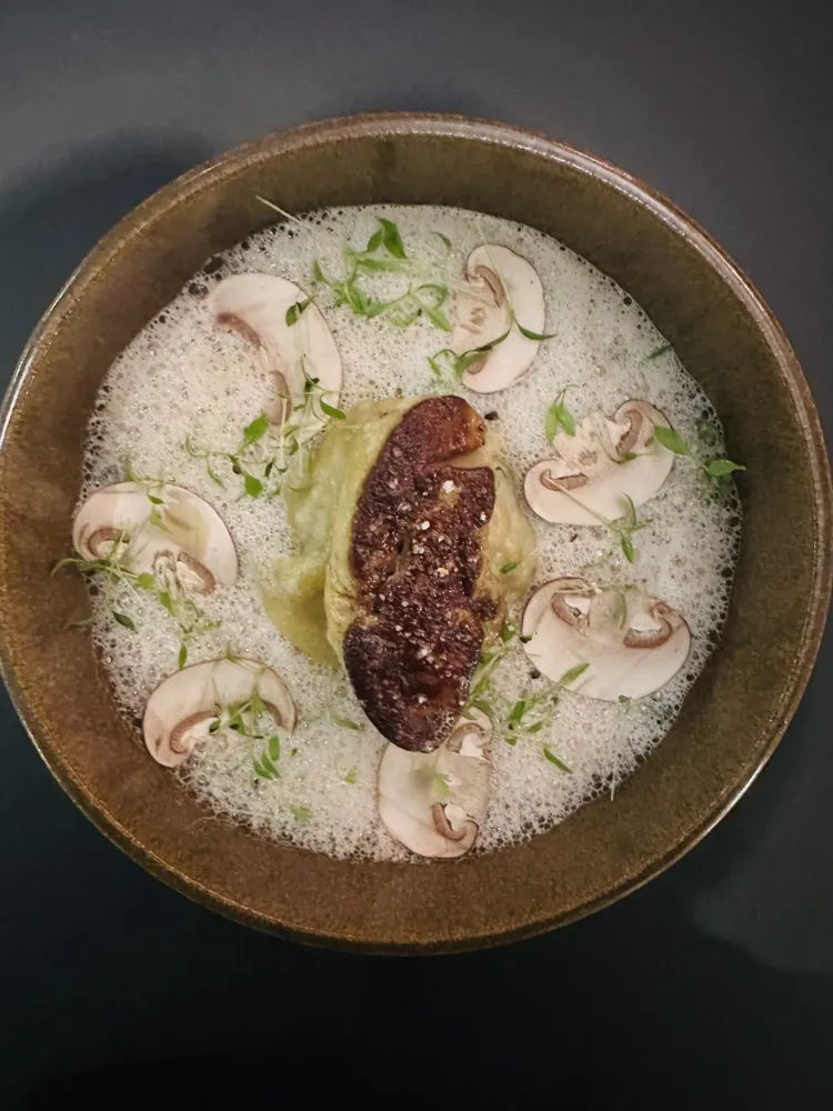 A bowl containing soup with mushroom slices, sprouts, and a piece of black truffle
