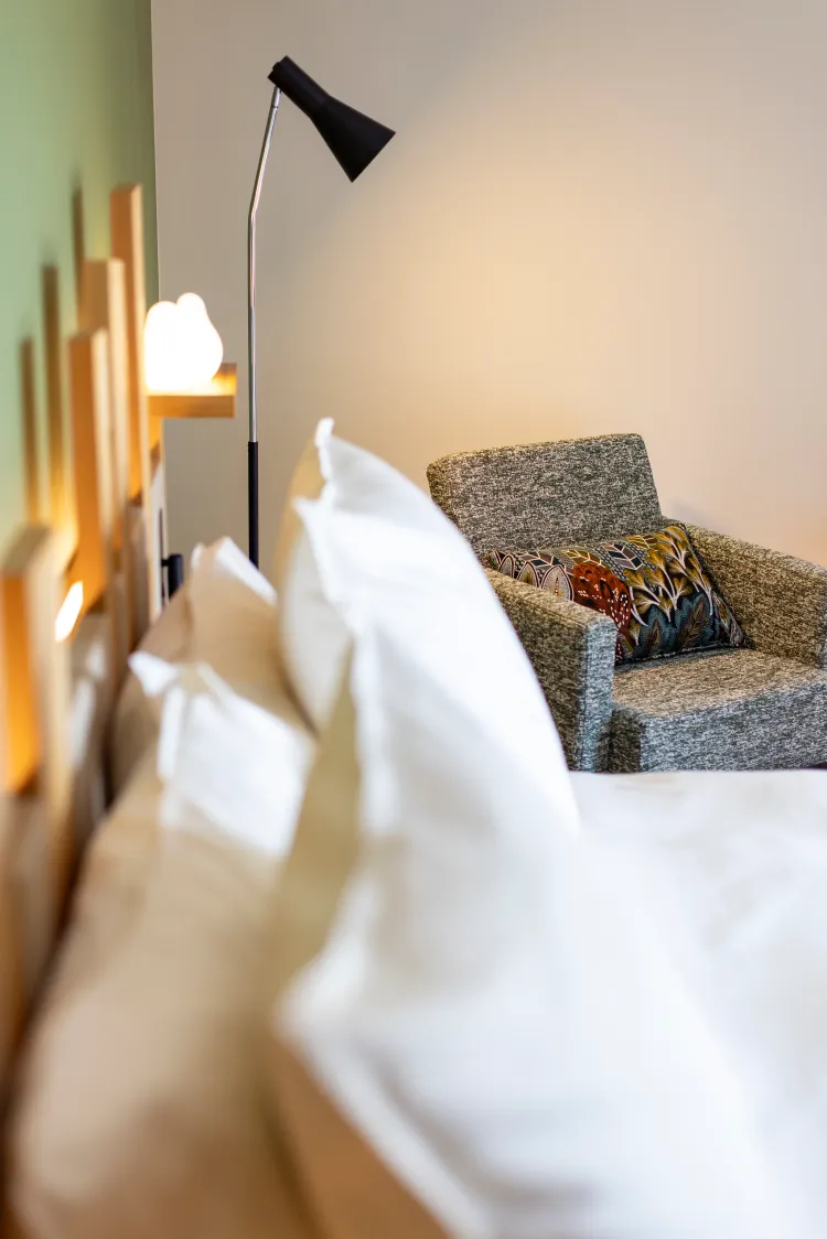 A bed with white pillows and a gray sofa with a decorative cushion, lit by a bedside lamp.