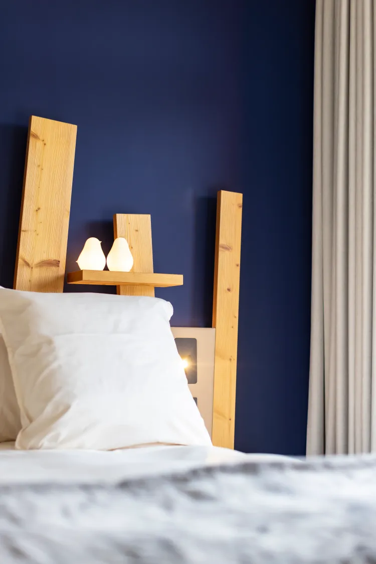 A bed with a light wood headboard and two lit candles placed on a wooden shelf, against a dark blue wall.