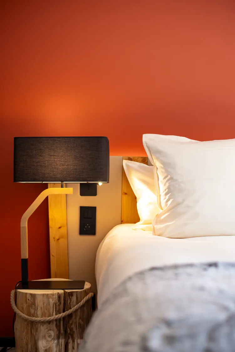 Bed with a light wood headboard, two white pillows, and a bedside lamp with a black shade on a raw wood nightstand.
