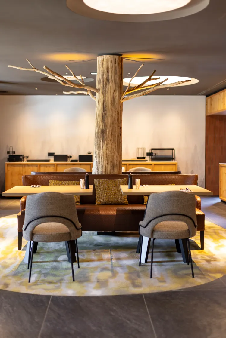 Ein Innenraum mit einem Tisch, der von Stühlen und einem Sofa umgeben ist, einem zentralen Baum mit beleuchteten Zweigen und einer Theke im Hintergrund