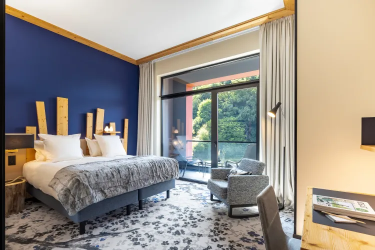 Zimmer mit einem Doppelbett, einem Nachttisch, einem Sessel und einer großen Fensterfront mit Zugang zu einer Terrasse mit Blick auf Bäume.